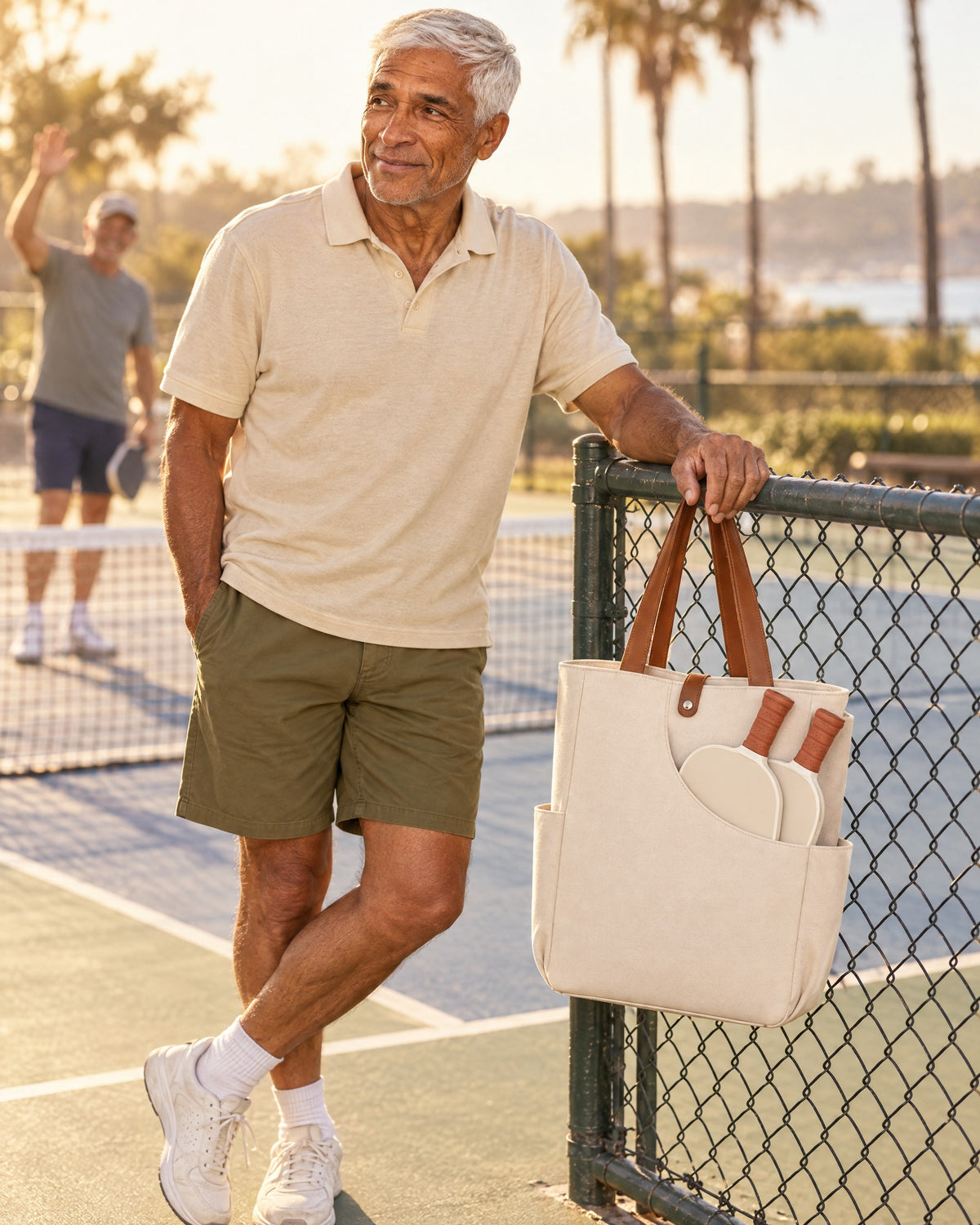 A man at the fence of a community pickleball court with the white Caleta Tote, two paddles in its front pocket, grips out