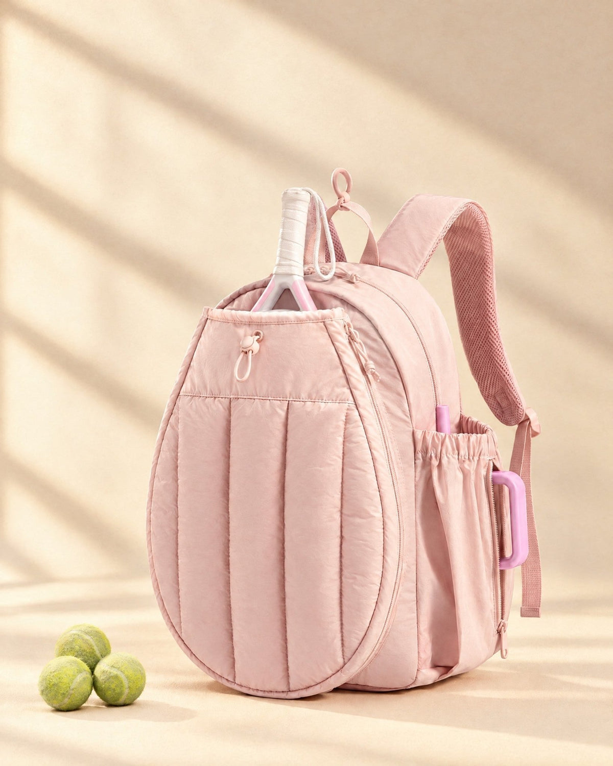 The Papagayo Backpack in pink, quilted, with a padel racket in its front pocket and tennis balls on the floor