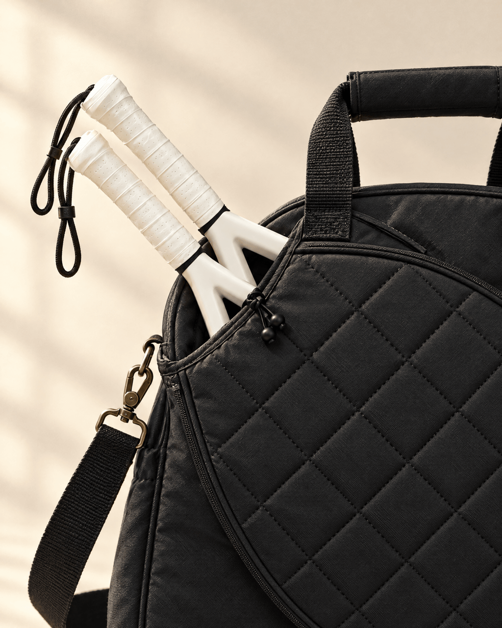 Close-up of the black diamond quilting, the toggle cord holding two padel racket handles, and the brass strap clip