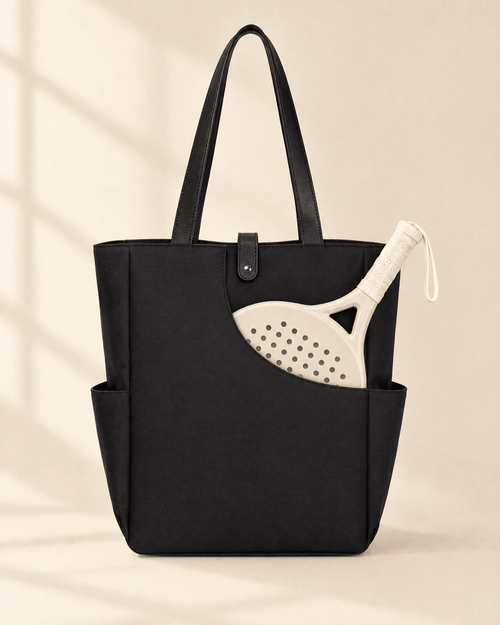 The Caleta Tote in black with black handles, a padel racket in its curved front pocket
