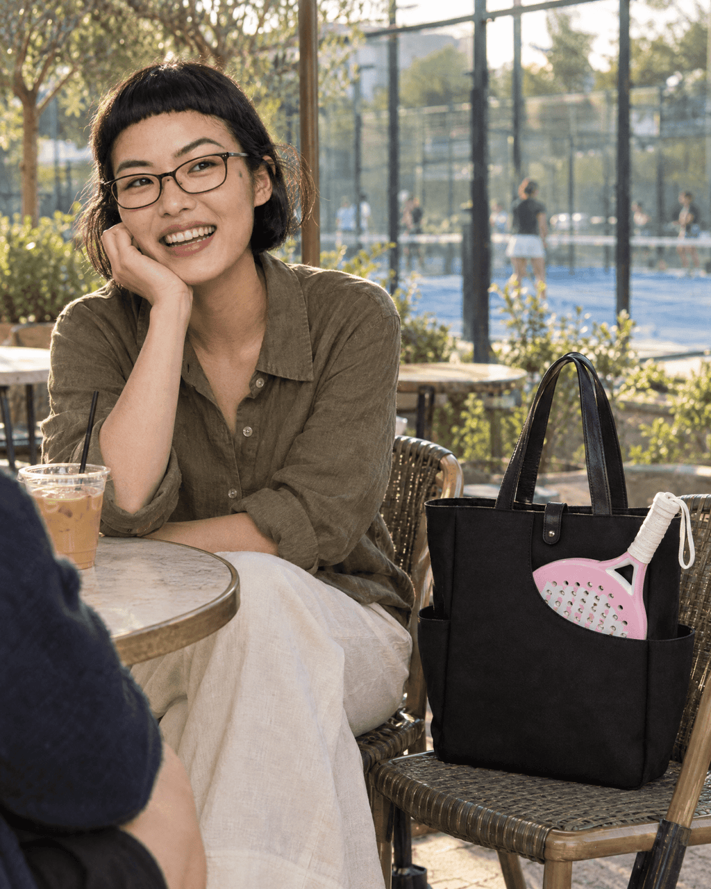 A woman laughing at a cafe beside a padel club, the black Caleta Tote on the chair next to her